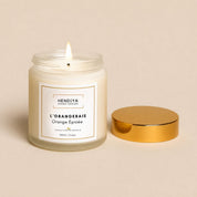 Bougie Végétale l'Orangeraie Hendiya en cire de soja végétale allumée, couvercle doré posé à côté sur fond beige, senteur orange épicée aux écorces de cannelier.
